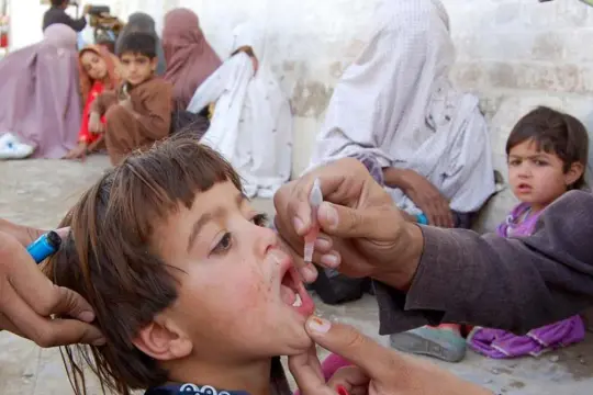 Dziecko otrzymuje krople przeciw polio. W tle inne dzieci i kobiety.