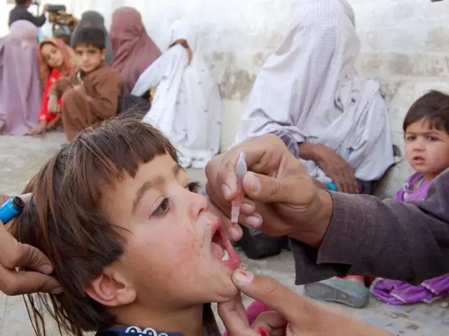 Dziecko otrzymuje krople przeciw polio. W tle inne dzieci i kobiety.