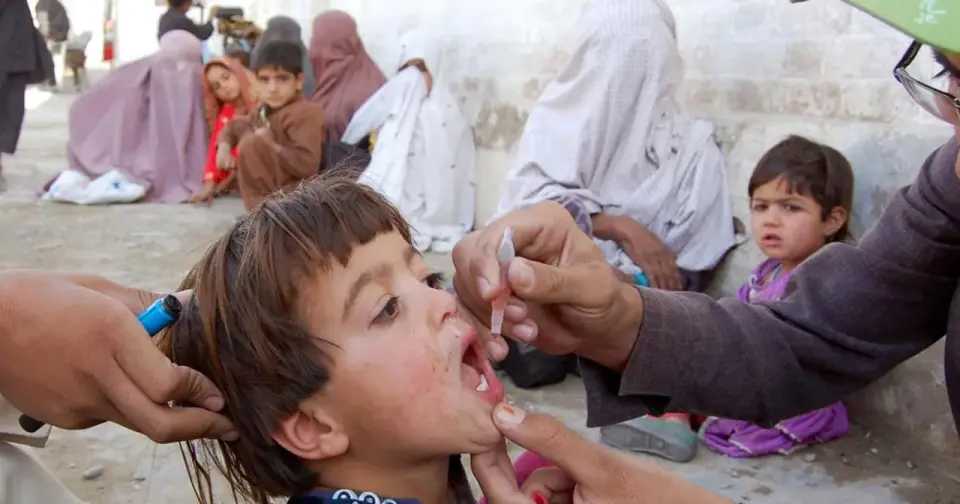 Dziecko otrzymuje krople przeciw polio. W tle inne dzieci i kobiety.